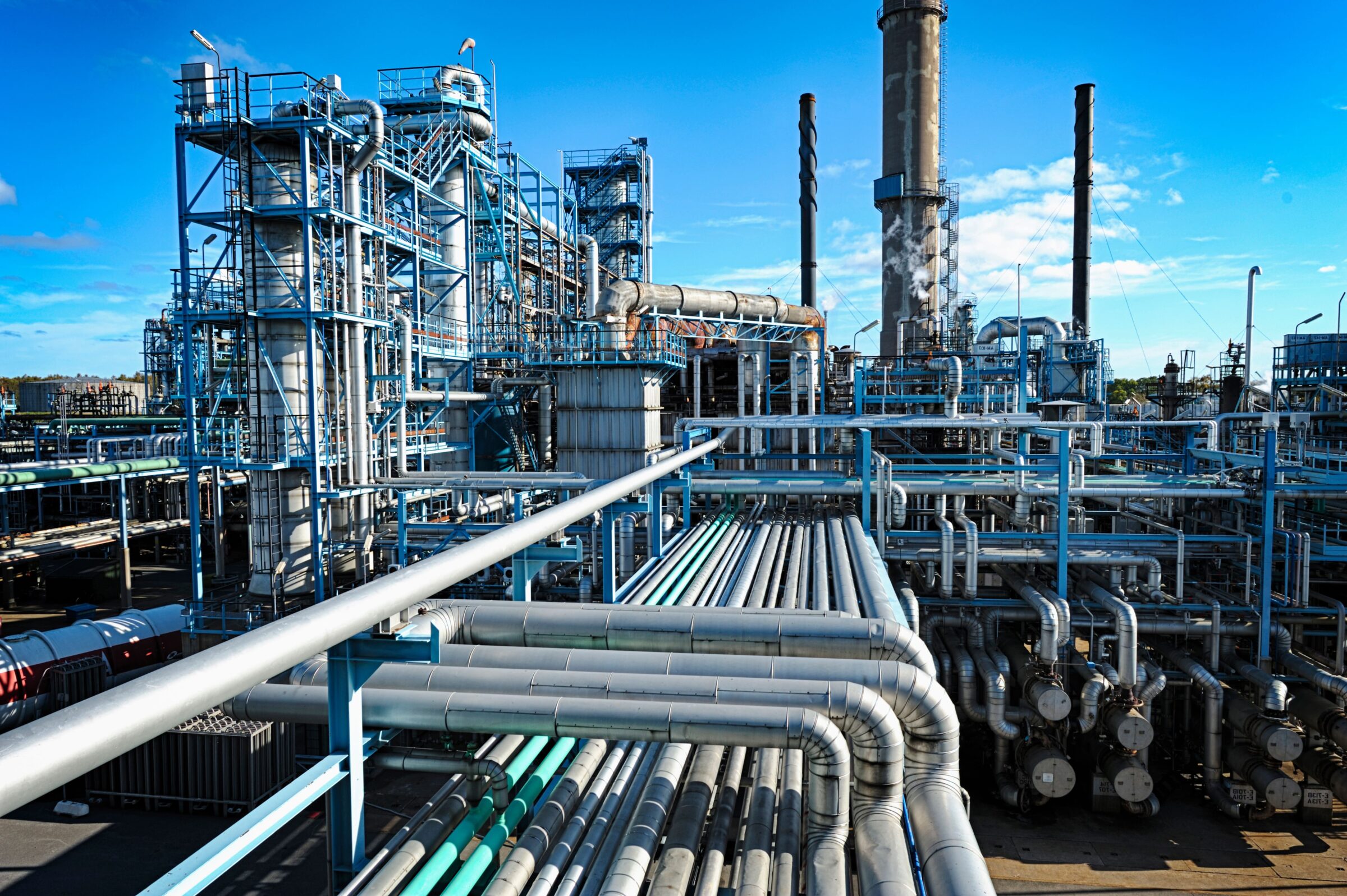 Industrial processing plant with extensive pipelines, steel structures, processing towers and chimneys under a clear blue sky.